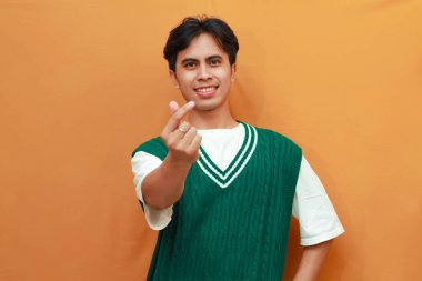 Yeşil kazaklı mutlu genç Asyalı adam ellerinde aşk işaretiyle poz veriyor ve turuncu arka planda izole edilmiş kameraya gülümsüyor.