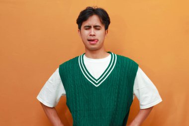 Yeşil kazaklı mutlu genç Asyalı adam turuncu arka planda izole edilmiş komik yüzlü komik bir poz veriyor.