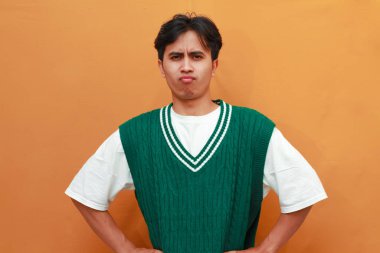 Yeşil kazaklı mutlu genç Asyalı adam turuncu arka planda izole edilmiş komik yüzlü komik bir poz veriyor.