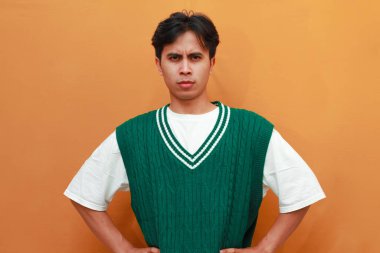 Asyalı genç adam yeşil kazak giyiyor. Kızgın yüz ve ellerle turuncu arka planda izole edilmiş kalçalarda poz veriyor.