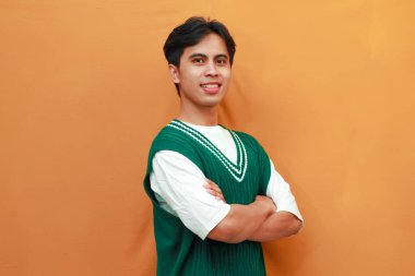 Gülümseyen ve kendinden emin yeşil kazaklı Asyalı genç adam kameraya çapraz kollarla bakıyor.