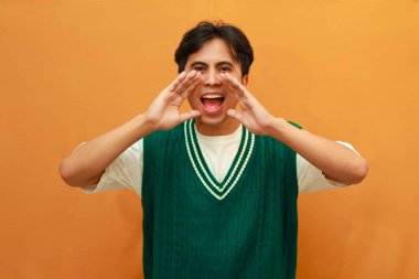 Yeşil kazaklı, gülümseyen ve kendinden emin Asyalı adam indirimli ya da turuncu arka planda izole edilmiş sıcak haberler sunuyor.