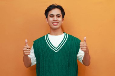 Yeşil kazaklı mutlu genç Asyalı adam turuncu arka planda parmaklarıyla işaret yapıyor.