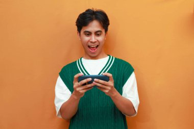 Yeşil kazaklı mutlu Asyalı adam oyun oynuyor ya da telefonda çığlıklar atıp zaferi ve başarıyı kutluyor.