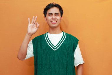 Yeşil kazaklı mutlu genç Asyalı adam turuncu arka planda parmaklarıyla işaret yapıyor.