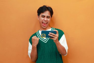 Yeşil kazaklı mutlu Asyalı adam oyun oynuyor ya da telefonda çığlıklar atıp zaferi ve başarıyı kutluyor.