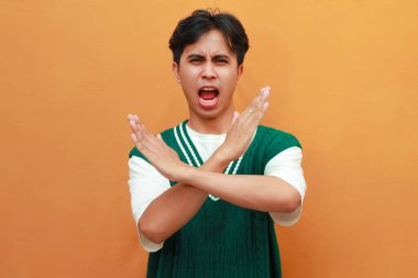 Ciddi bir yüz. Yeşil kazaklı genç Asyalı. Turuncu arka planda elle izole edilmiş dur işareti var.