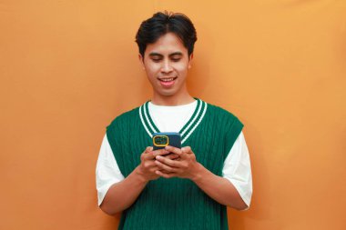 Mutlu yüz, yeşil kazaklı genç Asyalı adam. Odaklanıyor. Sosyal medyayı turuncu arka planda izole edilmiş cep telefonuna kaydırıyor.