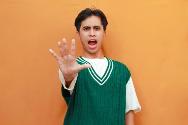 Ciddi bir yüz. Yeşil kazaklı genç Asyalı. Turuncu arka planda elle izole edilmiş dur işareti var.