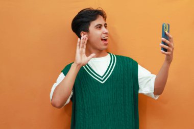 Yeşil kazak giyen mutlu genç Asyalı adam elinde akıllı telefonla poz veriyor ve turuncu arka planda izole edilmiş selfie çekiyor.