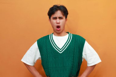 Ciddi bir yüz. Yeşil kazaklı genç Asyalı. Turuncu arka planda elle izole edilmiş dur işareti var.