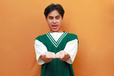 Yeşil kazaklı genç Asyalı adam her iki avucunun içi gibi mutlu, heyecanlı yüzünü turuncu arka planda gösteriyor.