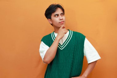 Yeşil kazaklı mutlu genç Asyalı adam turuncu arka planda izole edilmiş heyecanlı bir jest ile parlak bir fikir düşünüyor.