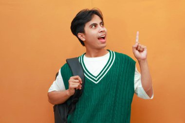 Yeşil kazaklı genç Asyalı öğrenci turuncu arka planda izole edilmiş parlak fikri düşünürken çenesini tutarak poz veriyor.