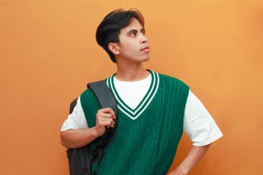 Yeşil kazaklı genç Asyalı öğrenci turuncu arka planda izole edilmiş parlak fikri düşünürken çenesini tutarak poz veriyor.