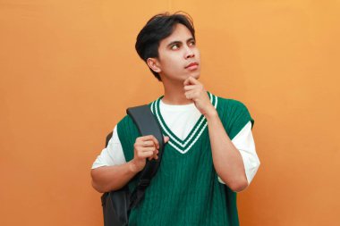 Yeşil kazaklı genç Asyalı öğrenci turuncu arka planda izole edilmiş parlak fikri düşünürken çenesini tutarak poz veriyor.