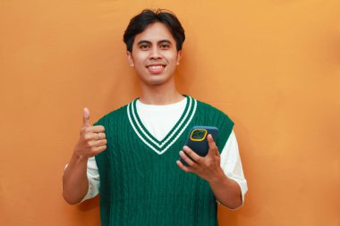 Yeşil kazaklı mutlu genç Asyalı adam elinde cep telefonuyla turuncu arka planda izole edilmiş güler yüzlü dostça bir jest işareti taşıyor.