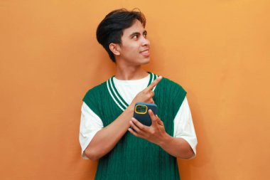 Yeşil kazaklı mutlu Asyalı adam cep telefonu tutuyor ve parmağıyla işaret ediyor.