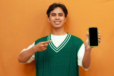 Yeşil kazaklı mutlu Asyalı adam cep telefonu tutuyor ve parmağıyla işaret ediyor.