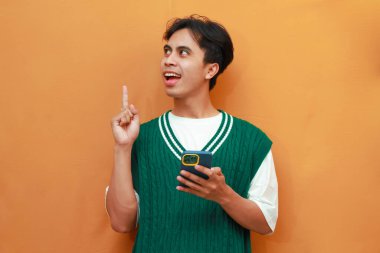 Yeşil kazaklı genç Asyalı adam elinde cep telefonuyla harika bir fikir olduğunu düşünüyor.