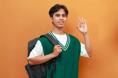 Yeşil kazaklı mutlu Asyalı öğrenci sırt çantası taşıyor ve turuncu arka planda parmaklarıyla tamam işareti yapıyor.
