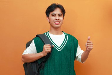 Yeşil kazaklı mutlu Asyalı öğrenci sırt çantası taşıyor ve turuncu arka planda parmaklarıyla tamam işareti yapıyor.