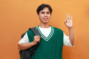 Yeşil kazaklı mutlu Asyalı öğrenci sırt çantası taşıyor ve turuncu arka planda parmaklarıyla tamam işareti yapıyor.