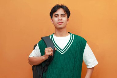 Yeşil süveter giyen mutlu Asyalı öğrenci sırt çantası taşırken ve başparmağıyla yan tarafı gösterirken turuncu arka planda dost canlısı sırıtıyor.