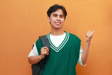 Yeşil süveter giyen mutlu Asyalı öğrenci sırt çantası taşırken ve başparmağıyla yan tarafı gösterirken turuncu arka planda dost canlısı sırıtıyor.