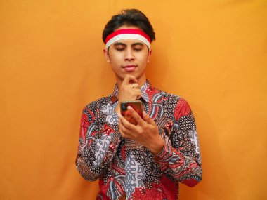 Batik tişörtlü ciddi Endonezyalı erkekler 17 Ağustos 'ta Endonezya Bağımsızlık Günü' nü kutluyor, cep telefonlarıyla poz veriyorlar ve el pençe divan sorular soruyorlar, sarı arka planda izole edilmiş kafa karıştırıcı fikirleri düşünüyorlar..