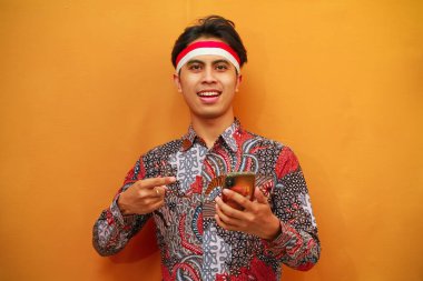 Batik gömlekli gülümseyen Endonezyalı erkekler, 17 Ağustos 'ta Endonezya Bağımsızlık Günü' nü kutluyor, ellerinde cep telefonları ve sarı arka planda izole edilmiş parmaklarıyla işaret ediyorlar..