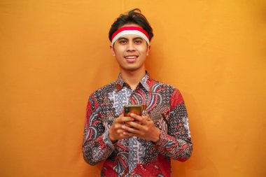 Batik gömlekli gülümseyen Endonezyalı erkekler, 17 Ağustos 'ta Endonezya Bağımsızlık Günü' nü kutluyor, ellerinde cep telefonları ve sarı arka planda izole edilmiş parmaklarıyla işaret ediyorlar..