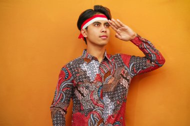 Batik tişörtlü Endonezyalı adam 17 Ağustos 'ta Endonezya Bağımsızlık Günü' nü kutluyor, elini kaldırıyor ve sarı arka planda izole edilmiş bir şekilde selamlıyor..