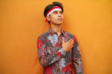 Batik tişörtlü Endonezyalı adam 17 Ağustos 'ta Endonezya Bağımsızlık Günü' nü kutluyor, elini kaldırıyor ve sarı arka planda izole edilmiş bir şekilde selamlıyor..