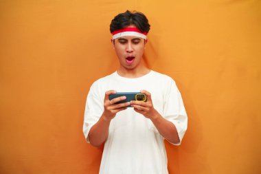 Mutlu ve heyecanlı Endonezyalı adam akıllı telefon oyunu oynuyor veya sarı arka planda dijital video akışı izliyor. 17 Ağustos 'ta Endonezya' nın bağımsızlık gününü kutlamak için özel reklam amaçlı mükemmel..
