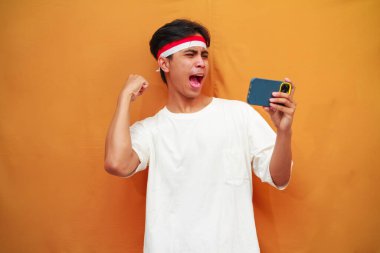 Mutlu ve heyecanlı Endonezyalı adam akıllı telefon oyunu oynuyor veya sarı arka planda dijital video akışı izliyor. 17 Ağustos 'ta Endonezya' nın bağımsızlık gününü kutlamak için özel reklam amaçlı mükemmel..