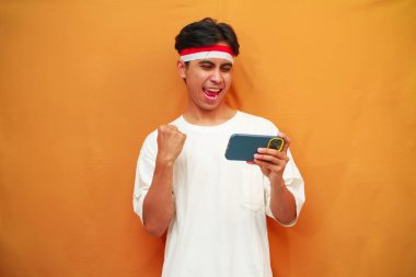 Mutlu ve heyecanlı Endonezyalı adam akıllı telefon oyunu oynuyor veya sarı arka planda dijital video akışı izliyor. 17 Ağustos 'ta Endonezya' nın bağımsızlık gününü kutlamak için özel reklam amaçlı mükemmel..