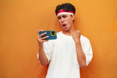 Mutlu ve heyecanlı Endonezyalı adam akıllı telefon oyunu oynuyor veya sarı arka planda dijital video akışı izliyor. 17 Ağustos 'ta Endonezya' nın bağımsızlık gününü kutlamak için özel reklam amaçlı mükemmel..