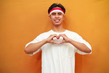 Gülümseyen Endonezyalı adam Endonezya Bağımsızlık Günü 'nü 17 Ağustos' ta kutluyor, elleriyle kalp işareti yapıyor. Sarı arka planda izole edilmiş Endonezya aşk sembolü.