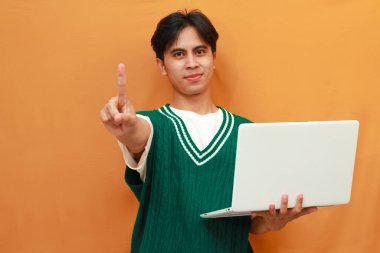 Yeşil kazaklı genç Asyalı adam elinde bilgisayarla gülümseyerek poz veriyor.