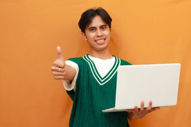 Yeşil kazaklı genç Asyalı adam dizüstü bilgisayarını tutuyor ve gülümseyen arkadaşça bir hareketle imza atıyor, mükemmel bir sembol.