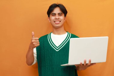 Yeşil kazaklı genç Asyalı adam dizüstü bilgisayarını tutuyor ve gülümseyen arkadaşça bir hareketle imza atıyor, mükemmel bir sembol.