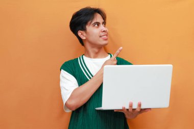 Yeşil kazak giyen neşeli genç Asyalı adam dizüstü bilgisayarını tutarken poz veriyor. Suratı asyalı adam dizüstü bilgisayarını turuncu arka planda tutarken poz veriyor.