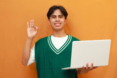 Yeşil kazaklı genç Asyalı adam dizüstü bilgisayarını tutuyor ve gülümseyen arkadaşça bir hareketle imza atıyor, mükemmel bir sembol.