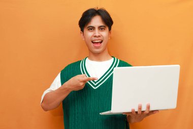 Yeşil kazak giyen neşeli genç Asyalı adam dizüstü bilgisayarını tutarken poz veriyor. Suratı asyalı adam dizüstü bilgisayarını turuncu arka planda tutarken poz veriyor.