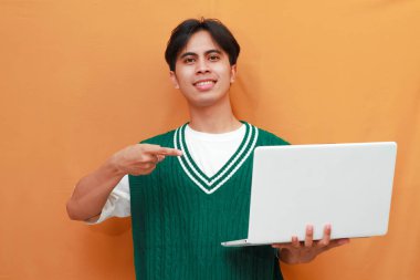 Yeşil kazak giyen neşeli genç Asyalı adam dizüstü bilgisayarını tutarken poz veriyor. Suratı asyalı adam dizüstü bilgisayarını turuncu arka planda tutarken poz veriyor.