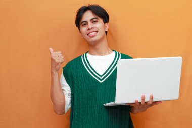 Yeşil kazak giyen neşeli genç Asyalı adam dizüstü bilgisayarını tutarken poz veriyor. Suratı asyalı adam dizüstü bilgisayarını turuncu arka planda tutarken poz veriyor.