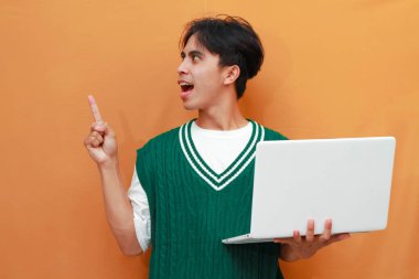 Yeşil kazak giyen neşeli genç Asyalı adam dizüstü bilgisayarını tutarken poz veriyor. Suratı asyalı adam dizüstü bilgisayarını turuncu arka planda tutarken poz veriyor.