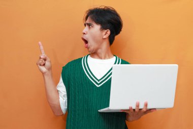 Yeşil kazak giyen neşeli genç Asyalı adam dizüstü bilgisayarını tutarken poz veriyor. Suratı asyalı adam dizüstü bilgisayarını turuncu arka planda tutarken poz veriyor.