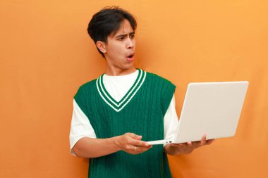 Yeşil kazaklı genç Asyalı adam elinde dizüstü bilgisayarla poz veriyor.